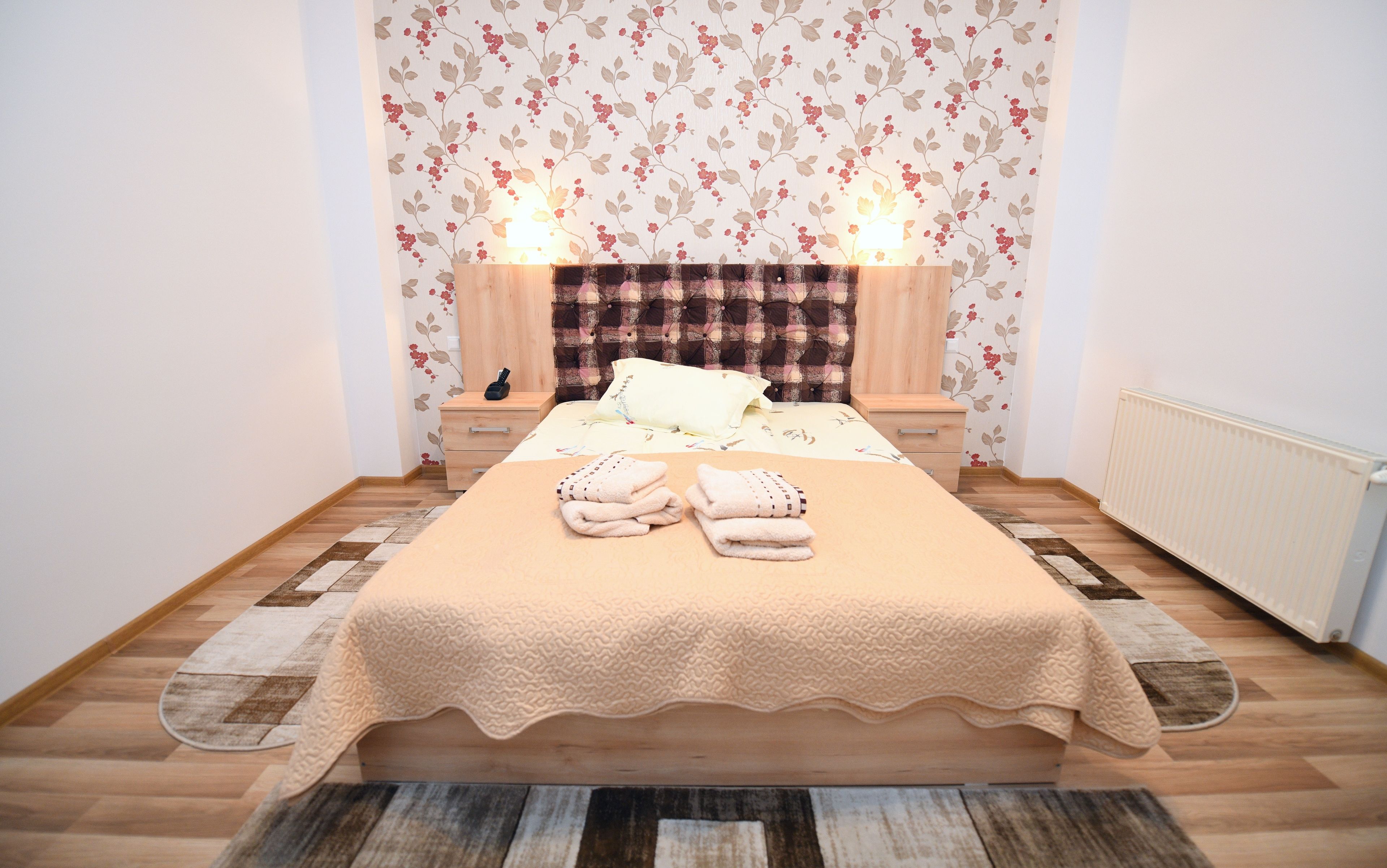 Un lit bien fait, recouvert d’une couverture beige, deux tables de chevet avec des lampes et un papier peint à motifs floraux.
