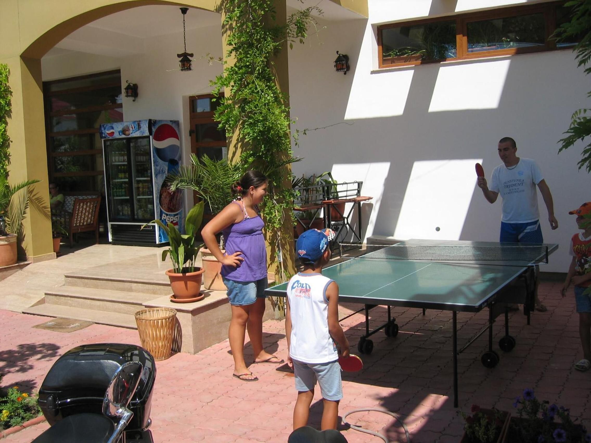 An outdoor ping pong table set up in a courtyard with potted plants and a Pepsi refrigerator in the background.