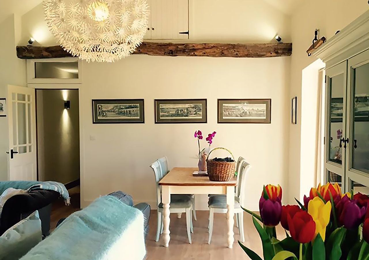 A dining area with a wooden table, white chairs, and a chandelier. There are framed pictures on the wall and a vase with flowers on the table.