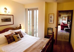 A bedroom with a large bed, a chair, and a door leading to a kitchen area. - Lackford Lakes Barns (Bury St Edmunds)