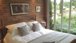 Una cama bien hecha con ropa blanca y almohadas estampadas, un cabecero de madera y vistas a la vegetación a través de amplias ventanas.