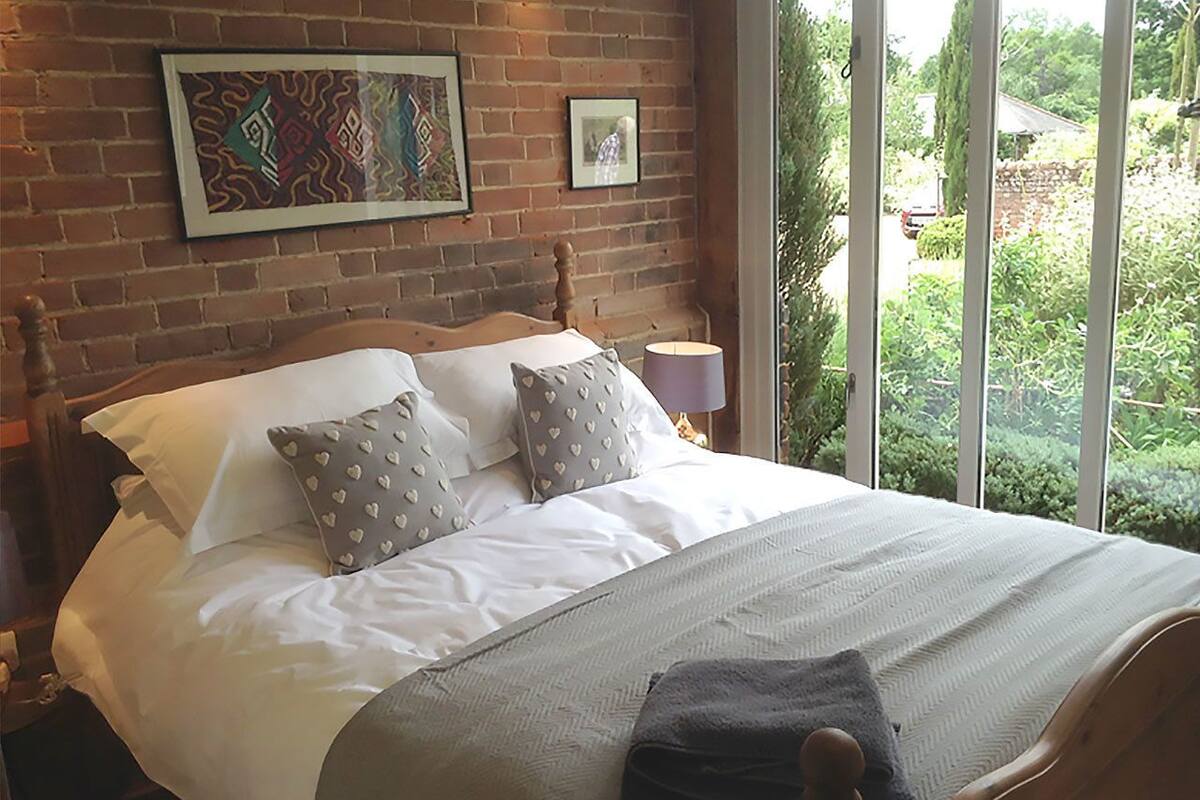 Un lit bien fait, avec du linge de lit blanc et des coussins à motifs, une tête de lit en bois et une vue sur la verdure à travers de grandes fenêtres.