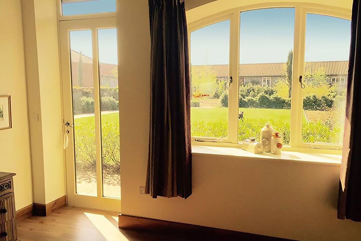 Une pièce dotée d’une grande porte-fenêtre coulissante donnant sur un jardin, d’une fenêtre avec des rideaux et d’une petite table sur laquelle sont posées des bouteilles.