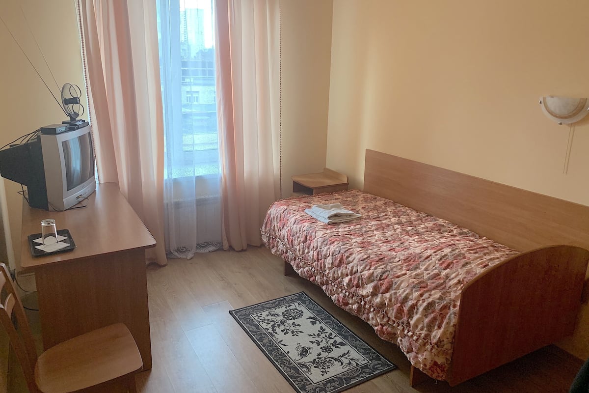 Een slaapkamer met een bed, een bureau met een televisie, een stoel, een nachtkastje en een raam met gordijnen.