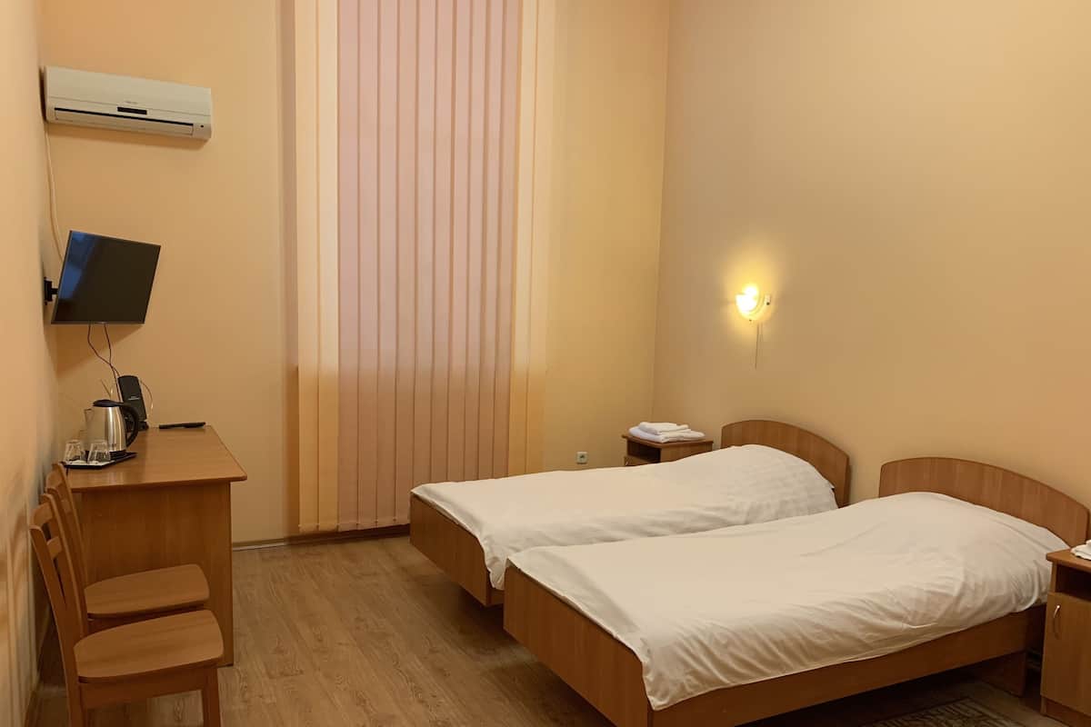 Een hotelkamer met twee houten bedden, een bureau met computer, een stoel, een nachtkastje met lamp en een raam met zonwering.