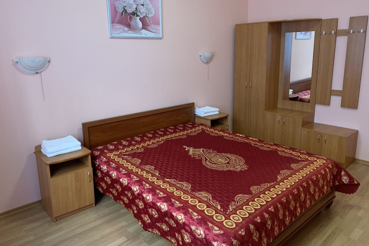 Een slaapkamer met een houten bed, een rode quilt met patroon, een houten kledingkast en een spiegel aan de muur.