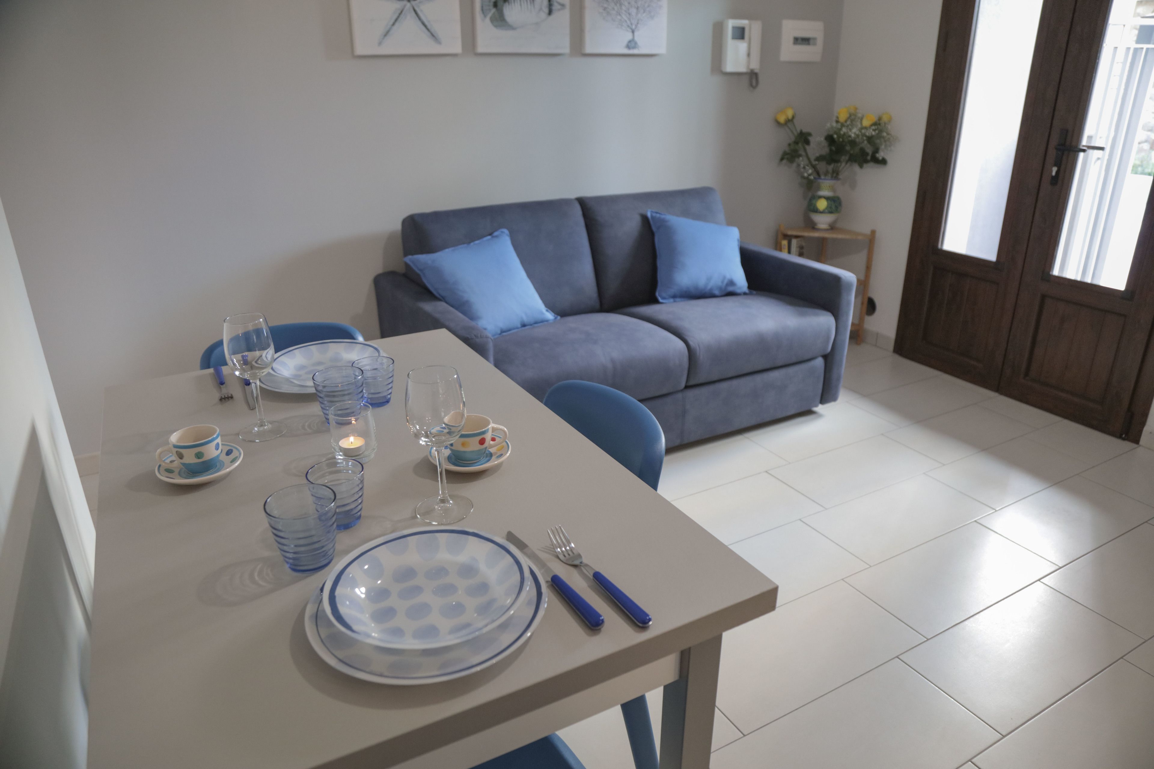 A dining area with a table set for two, a grey sofa, and a small side table with a vase of flowers.
