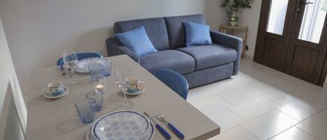 A dining area with a table set for two, a grey sofa, and a small side table with a vase of flowers.