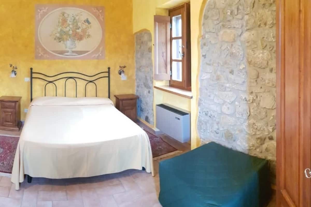 A bedroom with a bed, a nightstand, a dresser, a stone wall, and a window.