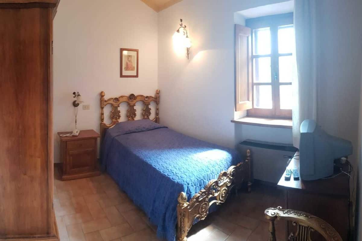 A bedroom with a wooden bed, a nightstand, a desk with a lamp, a window, and a picture on the wall.