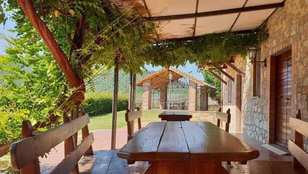 Un área de comedor al aire libre techada con mesa y bancos de madera, rodeada de muros de piedra y follaje.
