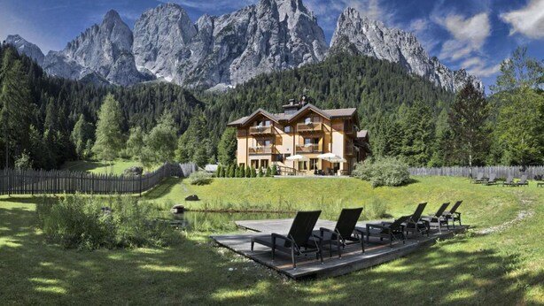 A chalet with a balcony, surrounded by a wooden deck and outdoor seating, set against a backdrop of mountains and forest.