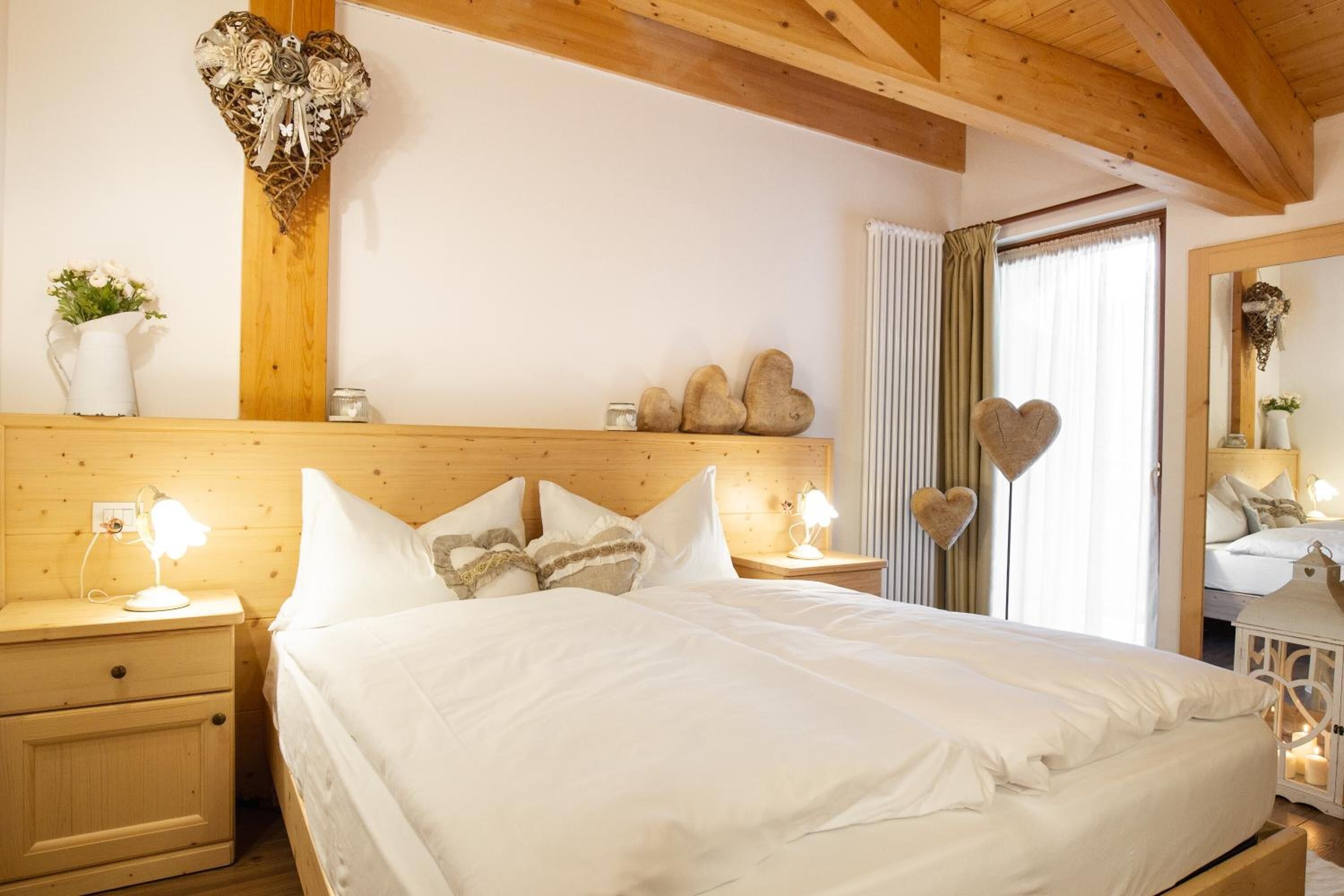 A bedroom with a wooden headboard, a bed with white bedding, two bedside tables with lamps, and decorative heart-shaped items on the wall and floor.