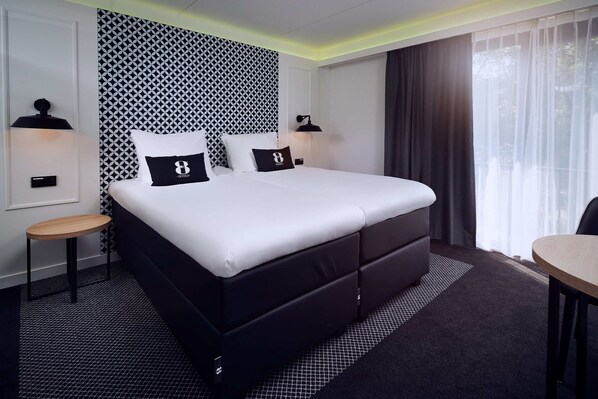 Una habitación de hotel moderna con una cama grande, dos mesitas de noche, un taburete de madera pequeño, un escritorio y un ventanal con cortinas traslúcidas.