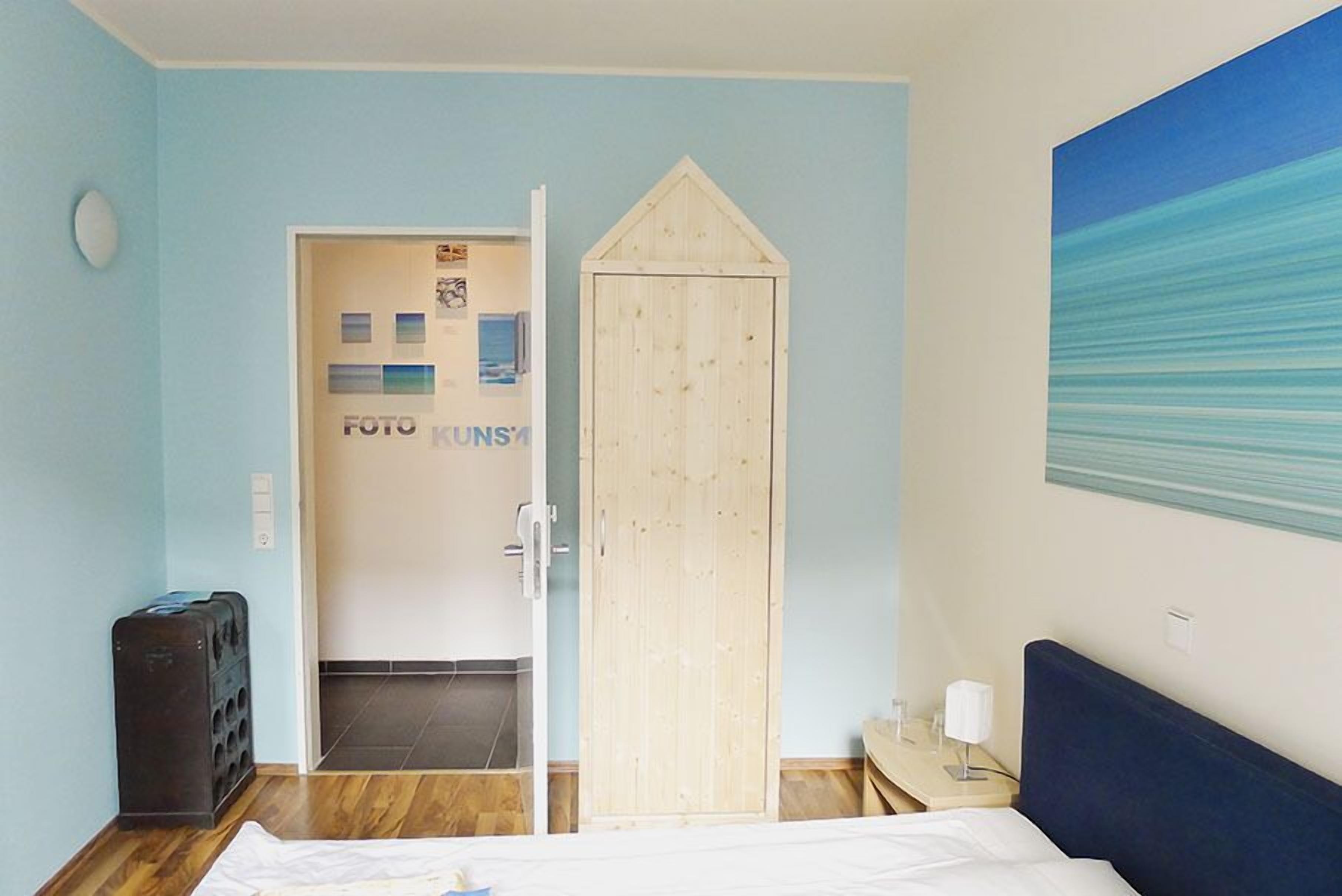 A hotel room with a wooden wardrobe, a bed, a small bedside table with a lamp, and a wall-mounted painting.