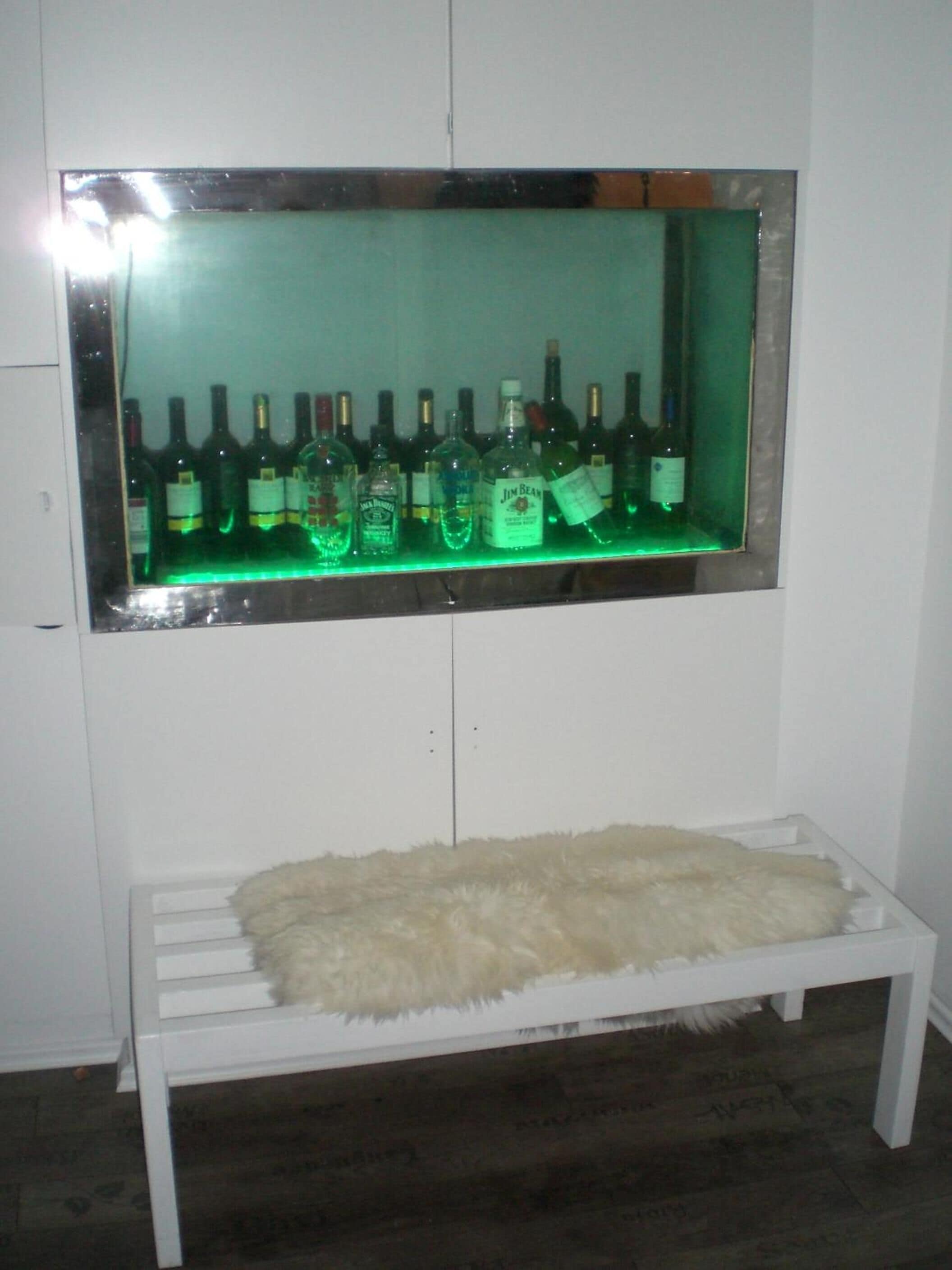 A bar area with a green-lit shelf displaying various liquor bottles. Below the shelf, there is a white bench with a fluffy white rug.