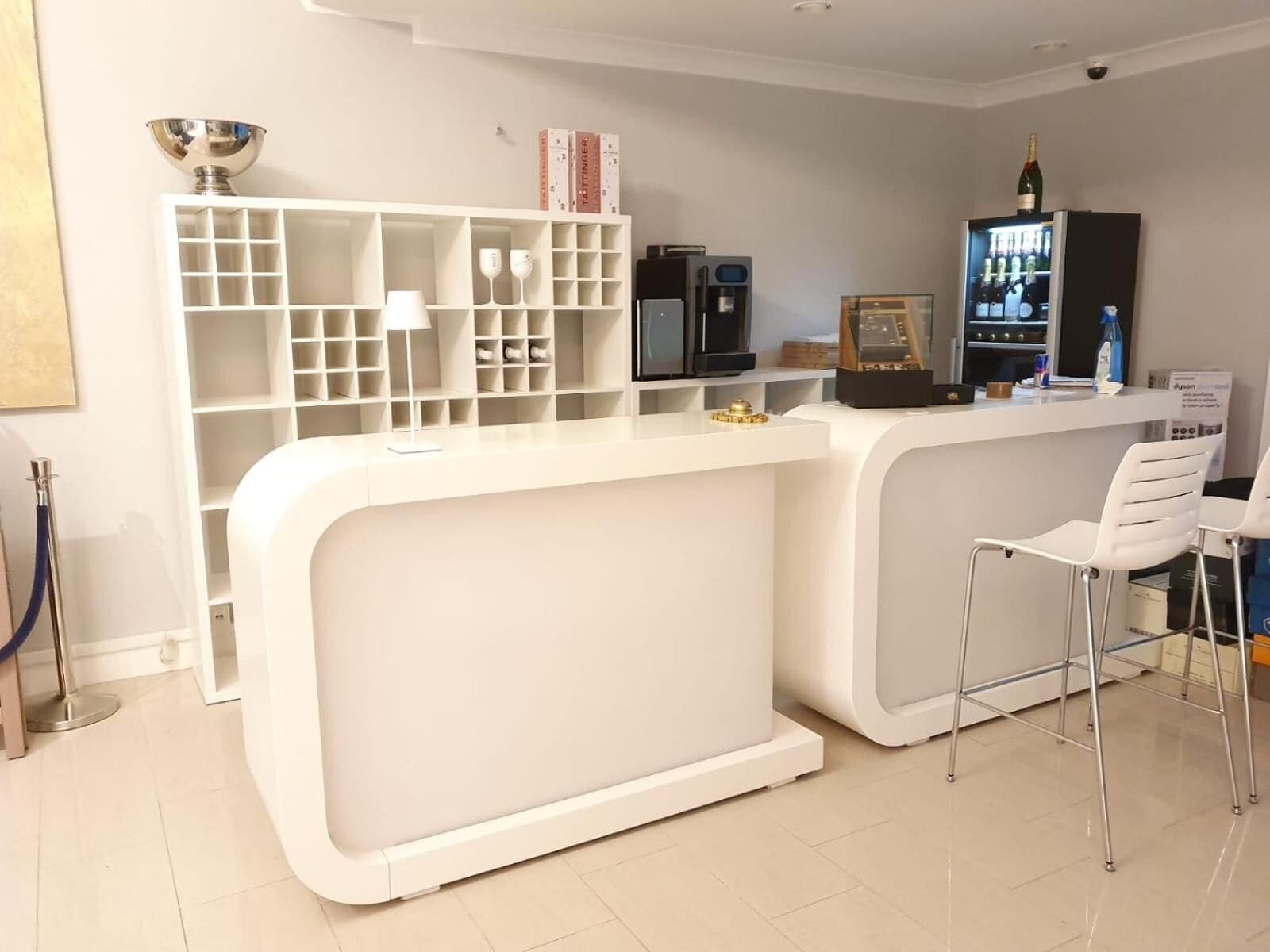 Een moderne, witte bar met een gebogen toonbank, een koffieapparaat en een minibars kastje. Er staan twee witte barstoelen en een plank met boeken en glazen.