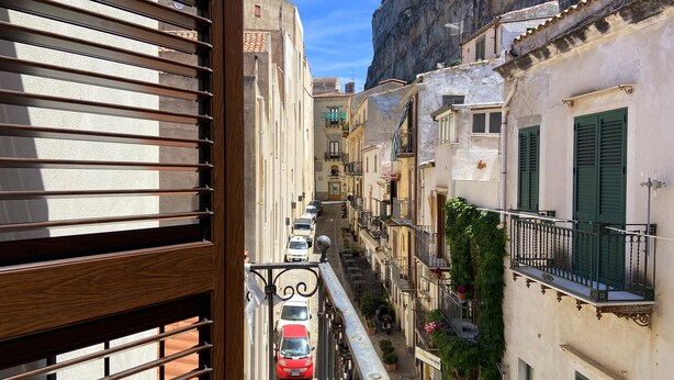 A narrow street with buildings on either side, a balcony with a view of parked cars, and a building with a sign that reads "duodo". - Terrazze XXV Novembre (Cefalù)