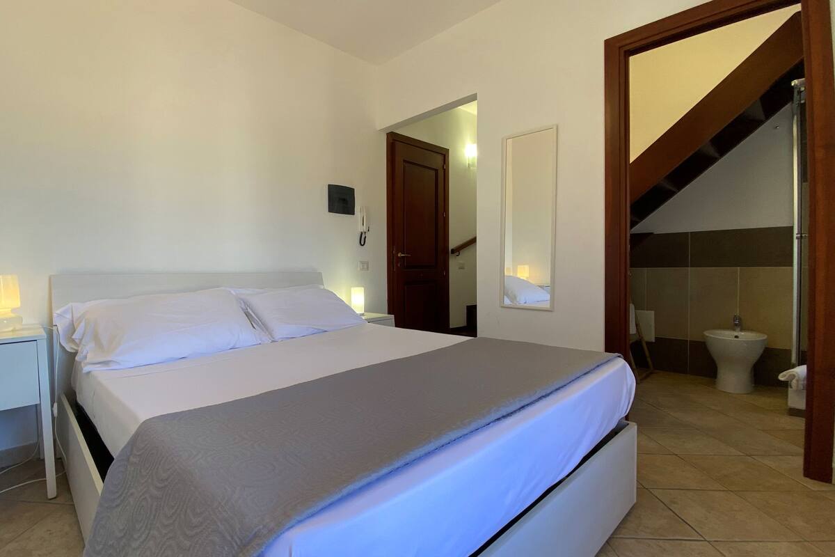 Un dormitorio ordenado con cama, mesitas de noche, un espejo y un baño visible a través de una puerta abierta.
