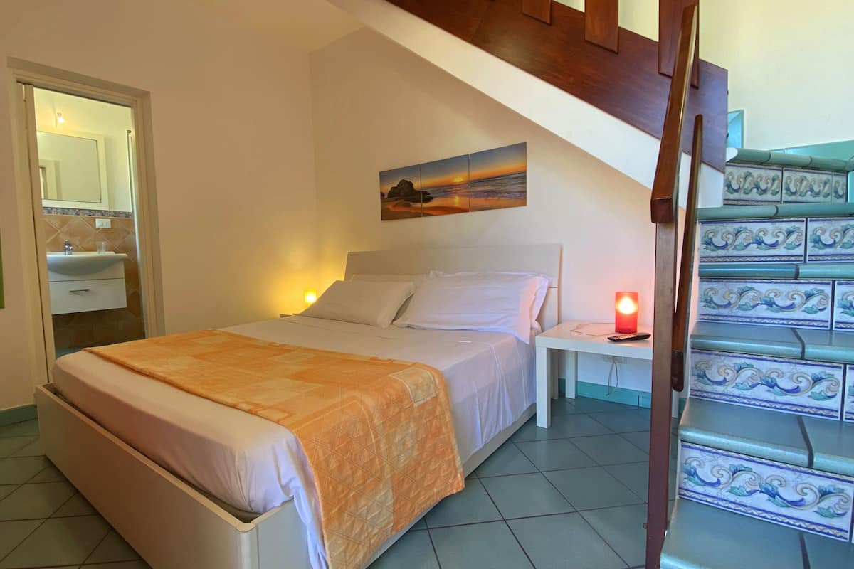 Un dormitorio con cama, una mesita de noche con lámpara, un cuadro, un baño con lavamanos y una escalera con azulejos decorativos.