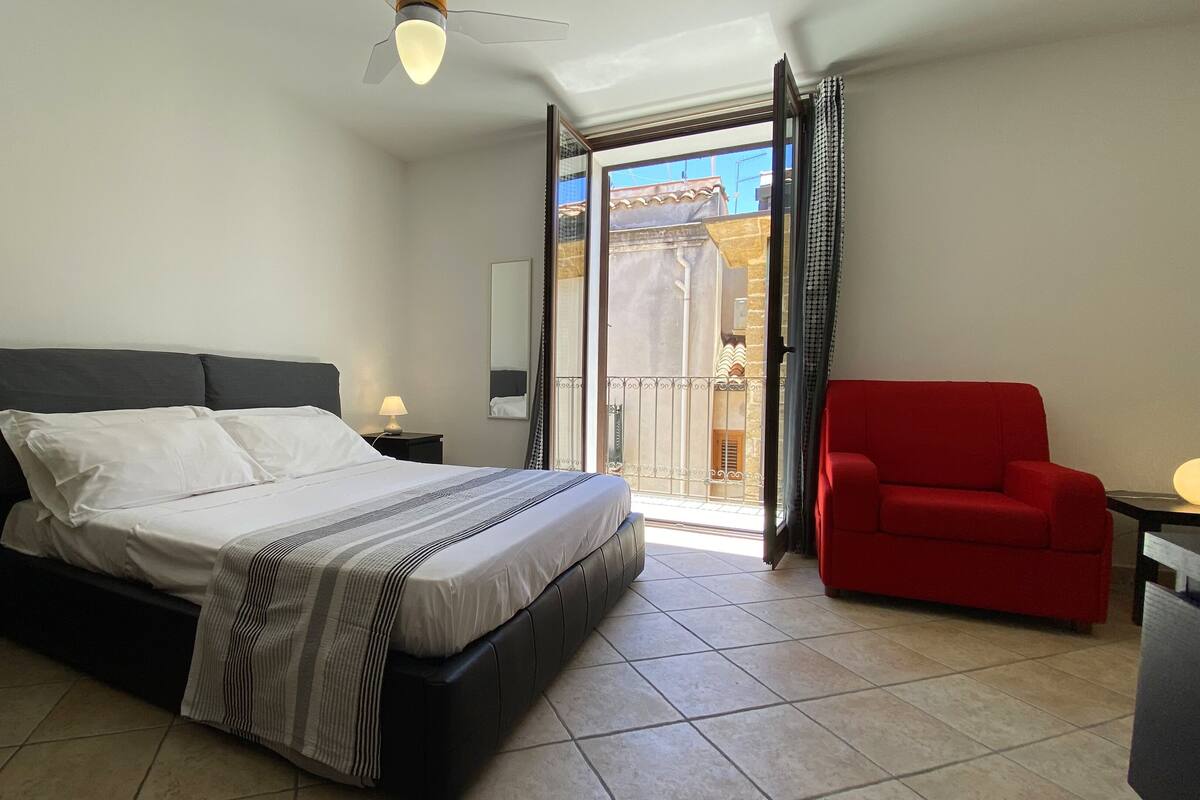 Un dormitorio con cama, mesita de noche, una silla roja, balcón con vistas y ventilador de techo.