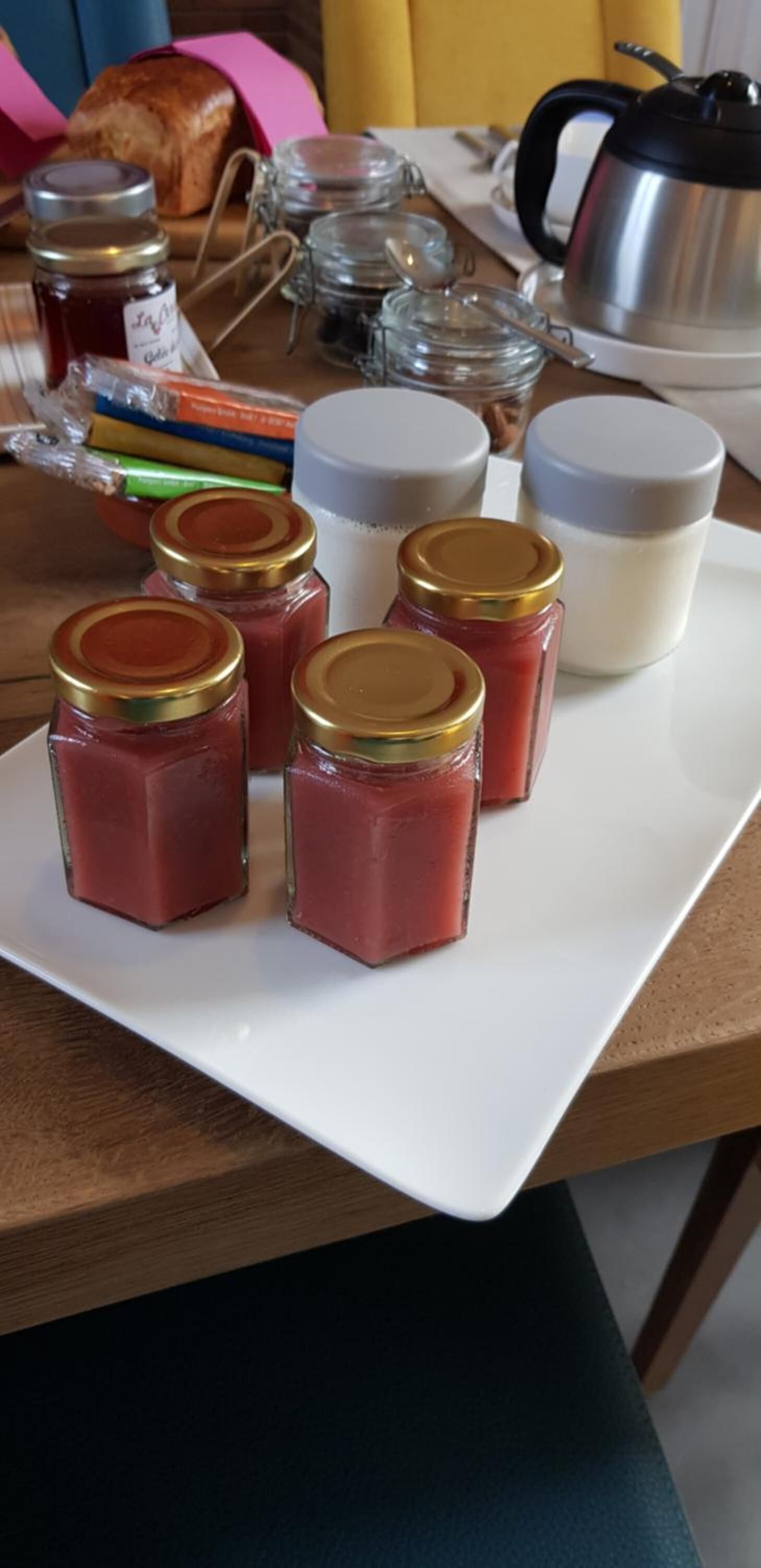 Un plateau blanc sur lequel on trouve des pots de confiture et de miel, une miche de pain, une cafetière et une tasse, le tout posé sur une table en bois.