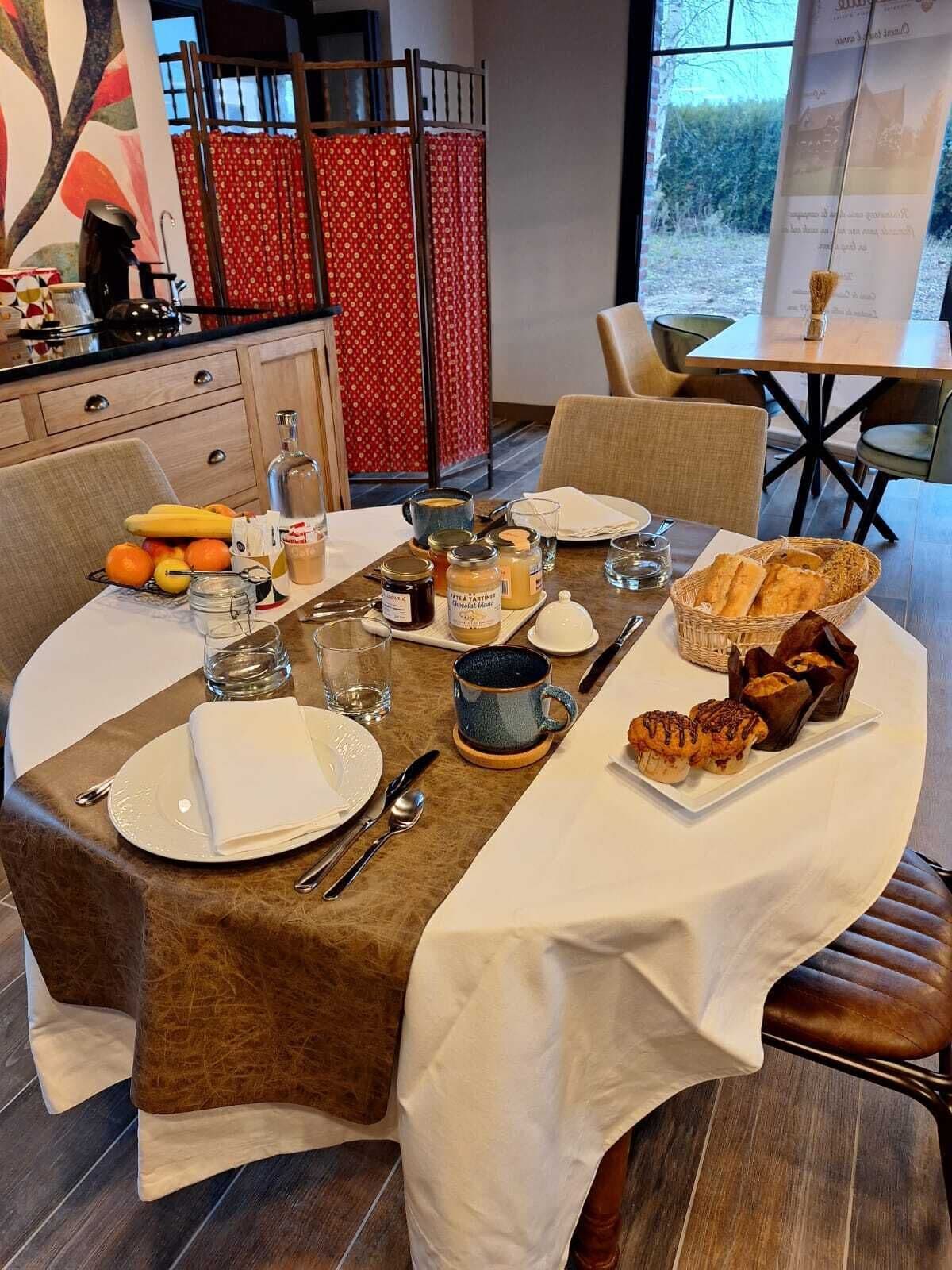 Una mesa de comedor dispuesta con platos, cubiertos, vasos y diversos alimentos, incluyendo pasteles y pan.