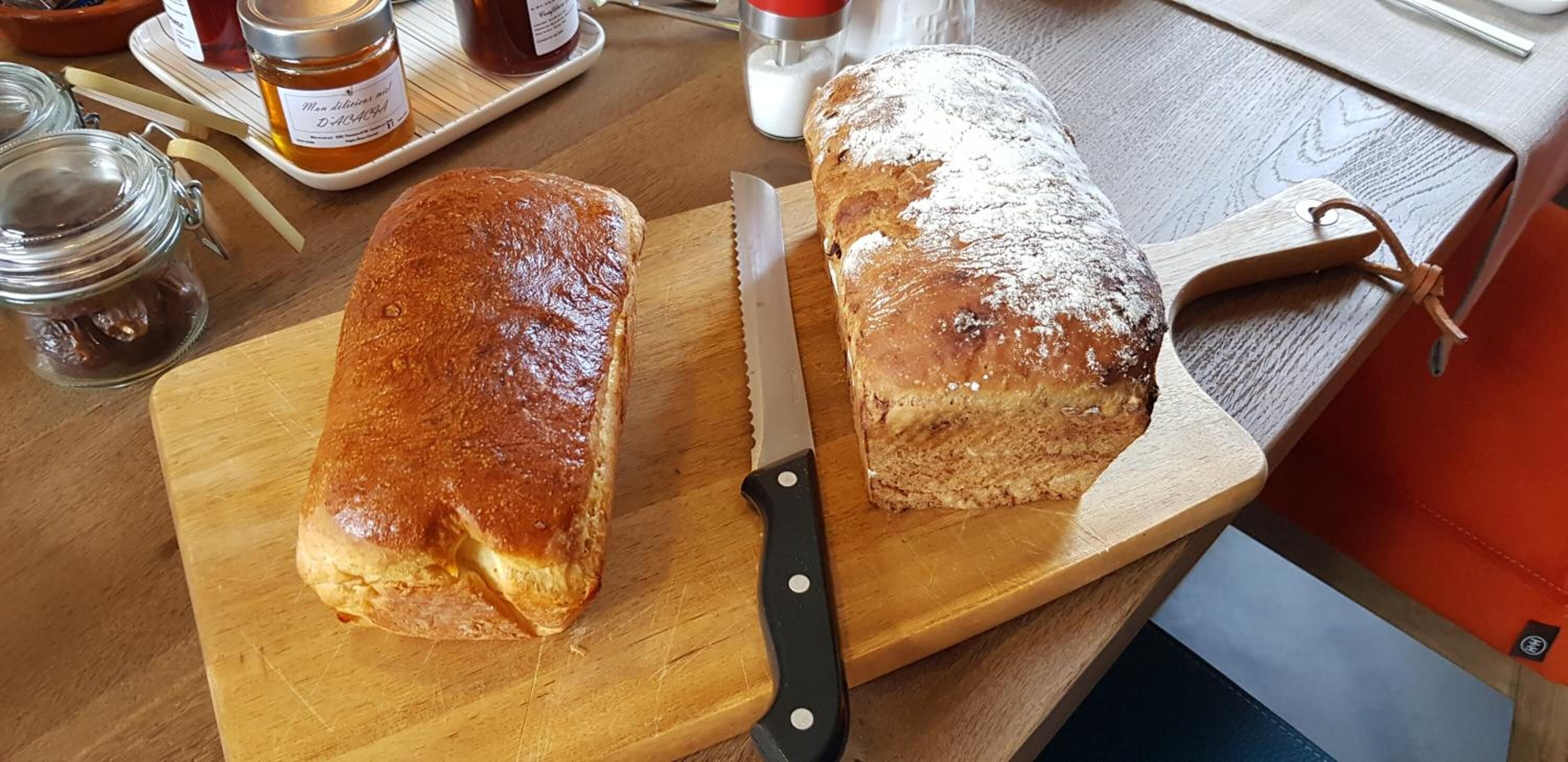 Dos hogazas de pan sobre una tabla de madera con un cuchillo, y tarros de miel al fondo.