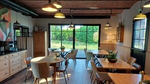 Un comedor amplio con mesas y sillas, ventanas grandes con vistas a zonas verdes, y un área de cocina con cafetera.