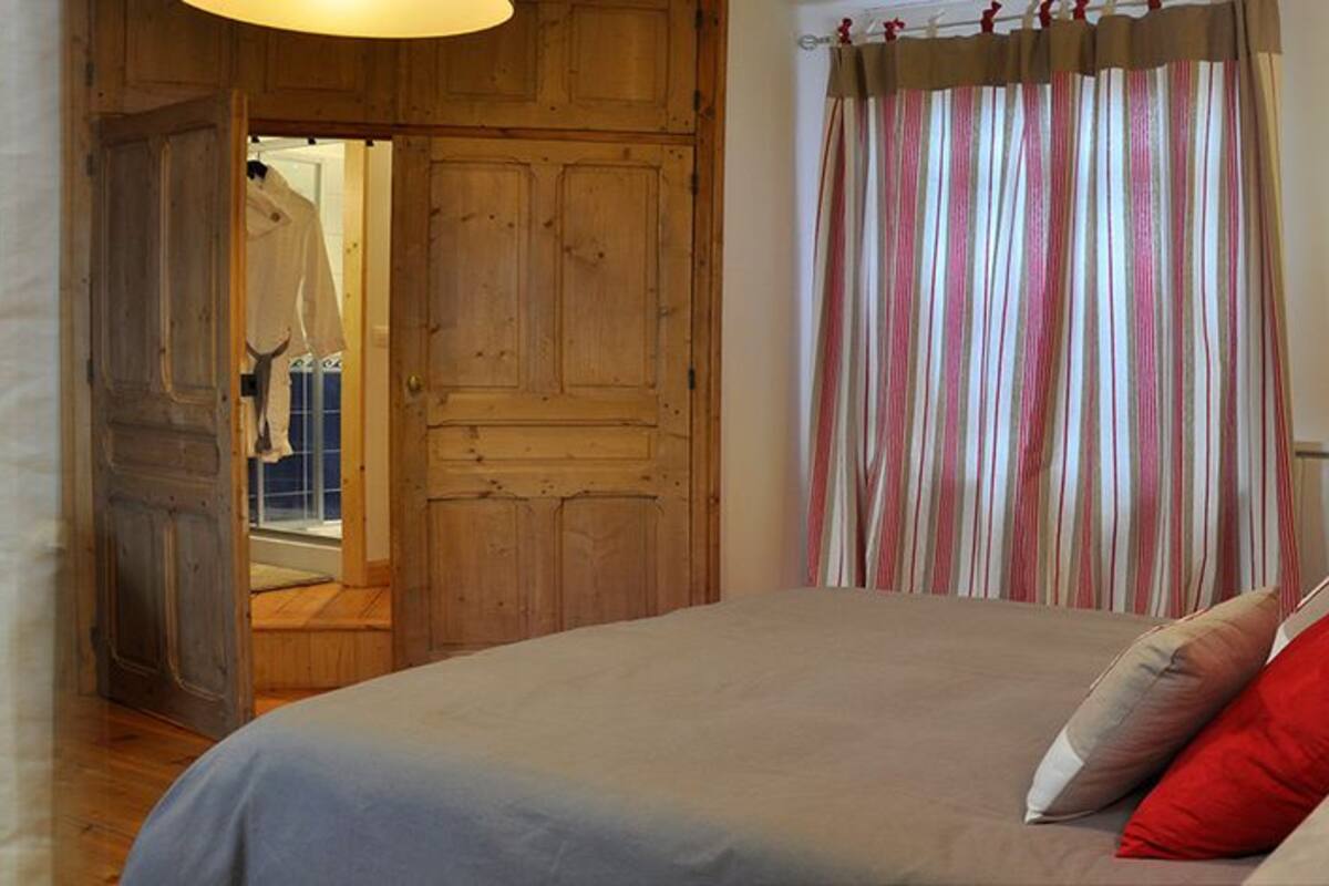 Un dormitorio con una cama, almohadas, una mesita de noche, una lámpara, un armario de madera y una cortina a rayas.