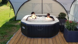 Un jacuzzi exterior con dos personas adentro, rodeado por una carpa y plantas en macetas.