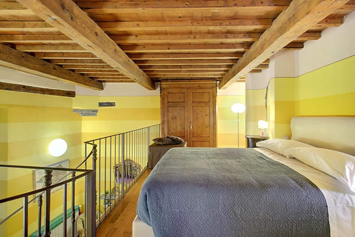 Une chambre avec un plafond en bois, un lit recouvert d’une couverture grise, une table de chevet avec une lampe et une porte en bois.