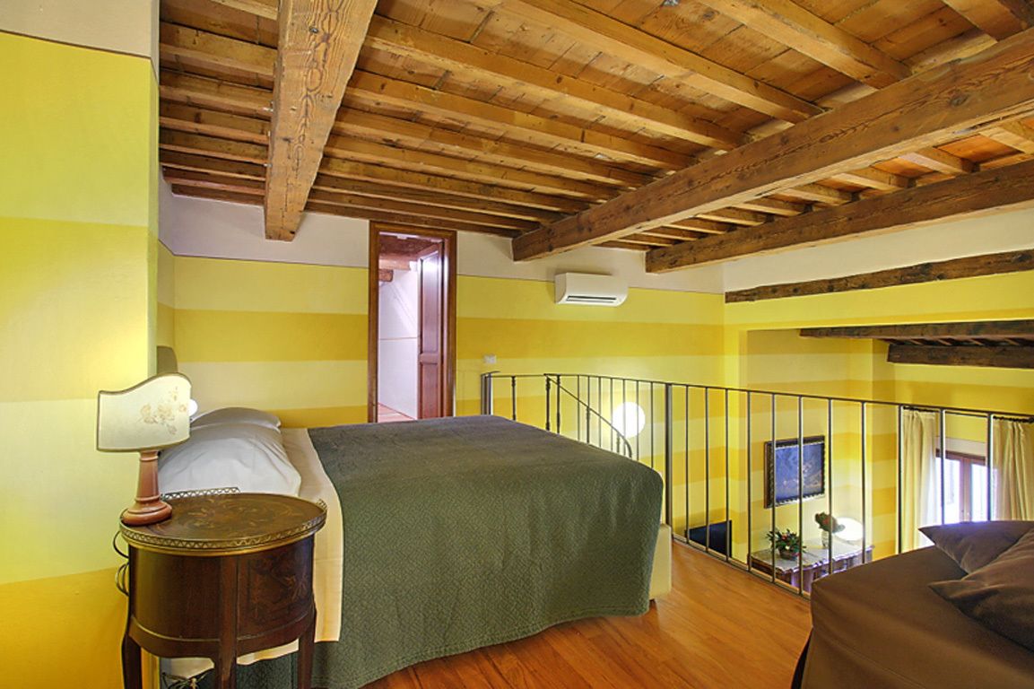 A bedroom with a wooden ceiling, a bed with a green bedspread, a small round table with a lamp, and a balcony with a railing.