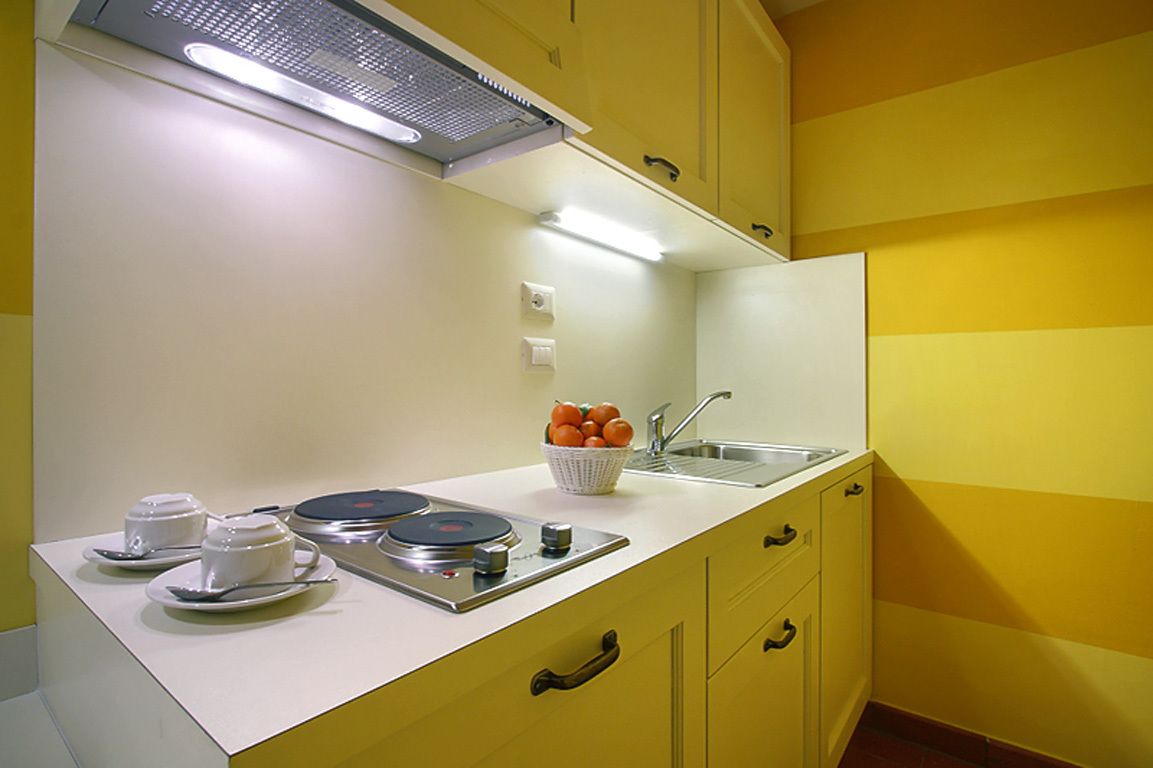 Une cuisine avec des meubles de couleur jaune, un plan de travail blanc, un évier et un saladier rempli de fruits.