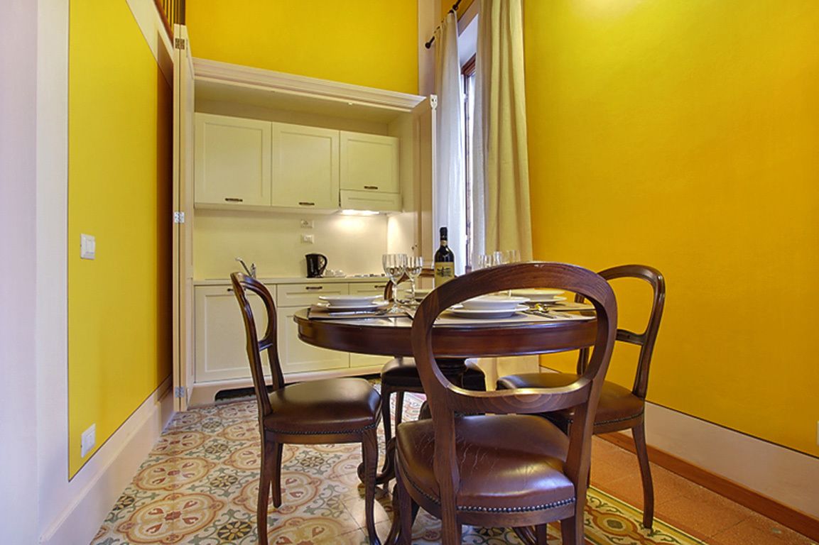 Un comedor con una mesa redonda y cuatro sillas, una cocina con armarios blancos y un suelo estampado.