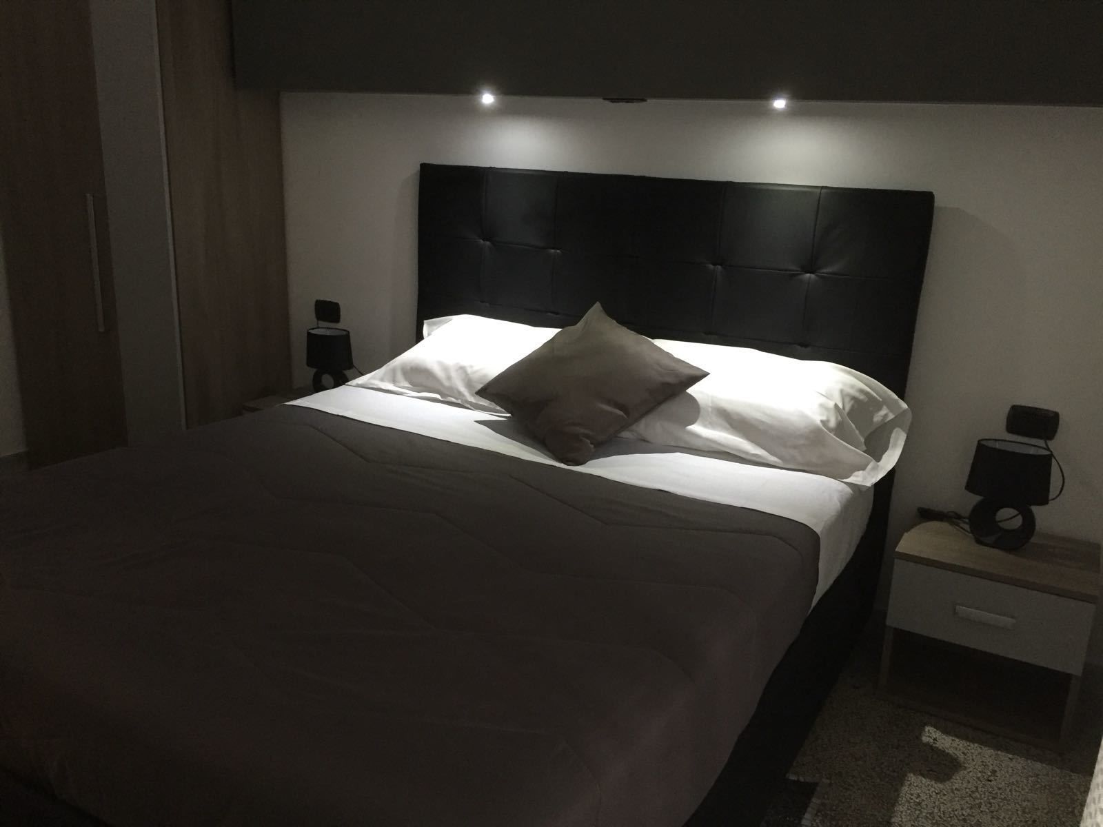 A neatly made bed with a black headboard, white linens, and a single pillow. There is a bedside table with a lamp and a small drawer.