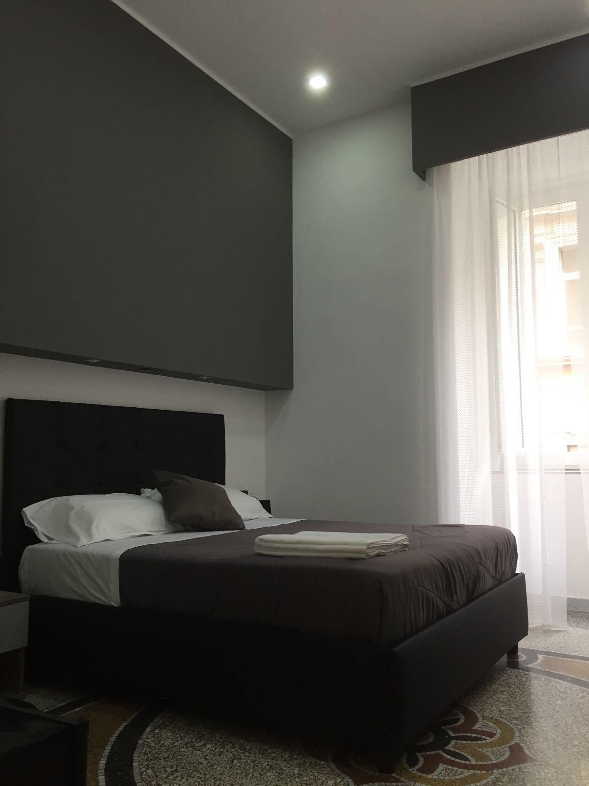 A modern hotel room with a large bed, a dark headboard, a small bedside table, and a large window with sheer curtains.