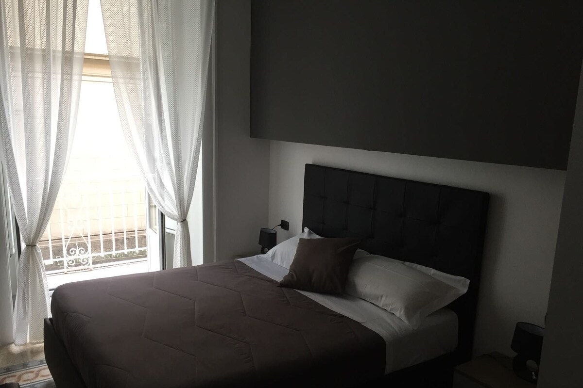 A bedroom with a large bed, a headboard, a bedside table, and a window with sheer curtains.