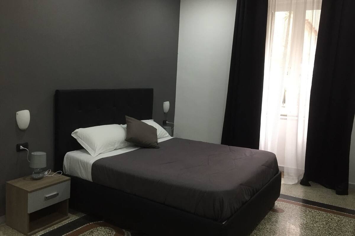 A modern bedroom with a large bed, a bedside table with a lamp, and a window with curtains.