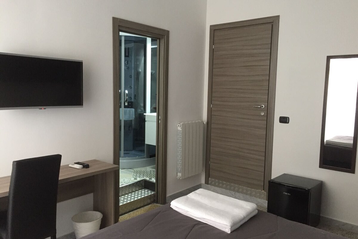 A hotel room with a bed, a desk, a chair, a TV, a wardrobe, and a small heater.
