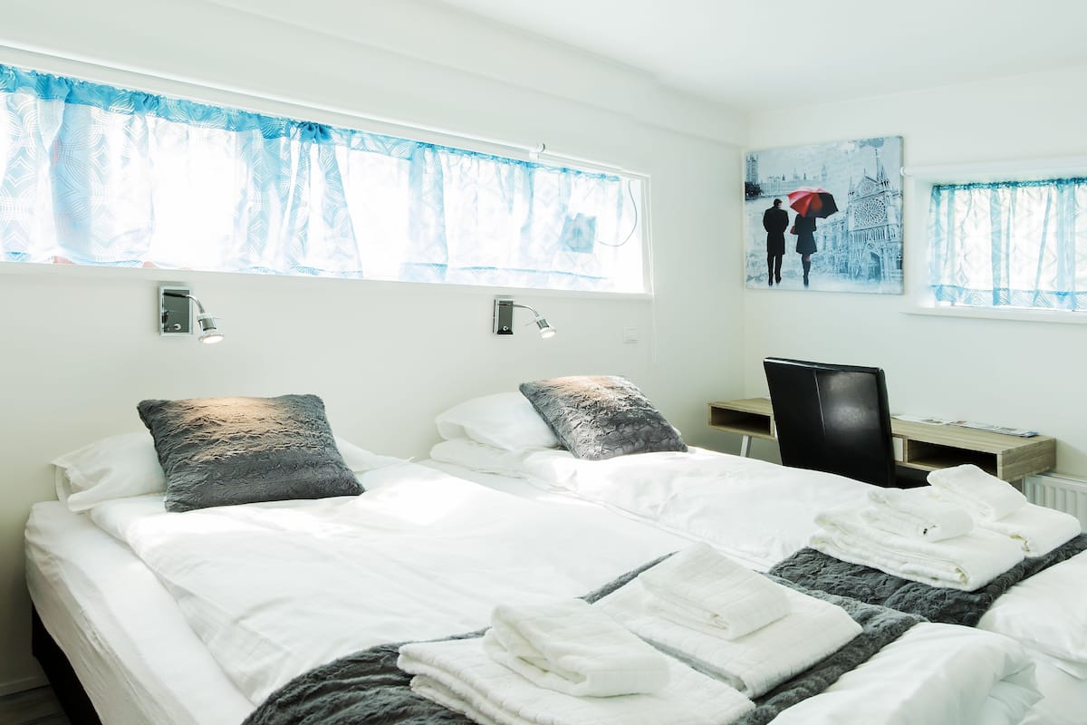 A hotel room with two beds, a desk, and a chair. There are two windows with curtains, a wall-mounted lamp, and a painting of a couple in a snowy landscape.