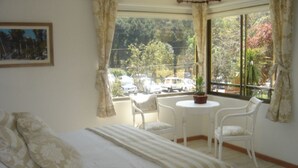 Un dormitorio con cama, una mesita redonda pequeña, dos sillas y una ventana con vistas a árboles y coches aparcados.