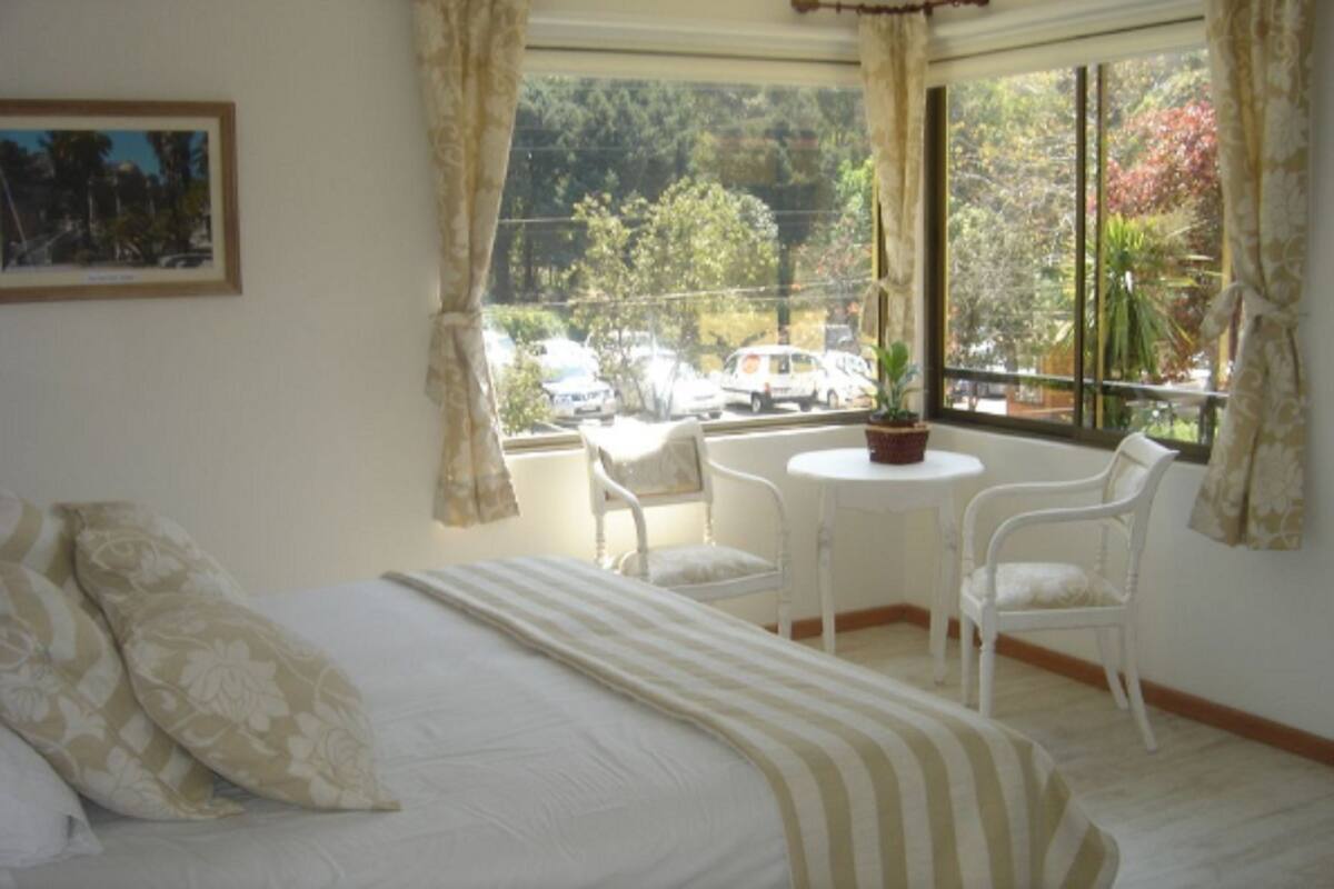 A bedroom with a bed, a small round table, two chairs, and a window with a view of trees and parked cars.