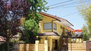 Una casa amarilla de dos pisos con una escalera que lleva al piso superior, un acceso con portón y un área exterior cubierta con una sombrilla.