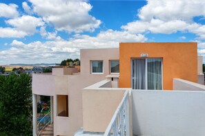 Ein mehrstöckiges Gebäude mit flachem Dach, einem Balkon mit weißer Geländer und Blick auf einen Spielplatz im Hintergrund.