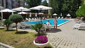 Ein Poolbereich mit Liegestühlen und Sonnenschirmen, umgeben von Gebäuden und viel Grün.