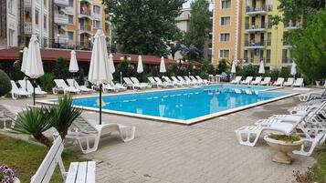 A swimming pool with sun loungers and umbrellas, surrounded by greenery and buildings.