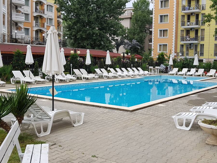 A swimming pool with sun loungers and umbrellas, surrounded by greenery and buildings.