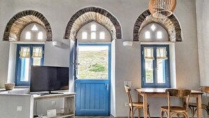 A room with a TV, a table, chairs, and a blue door leading to a scenic view. - Tinoshomelidays Residences (Tinos)