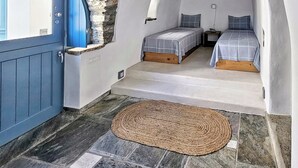 A bedroom with two beds, a stone wall, a wooden ceiling, and a blue door. - Tinoshomelidays Residences (Tinos)